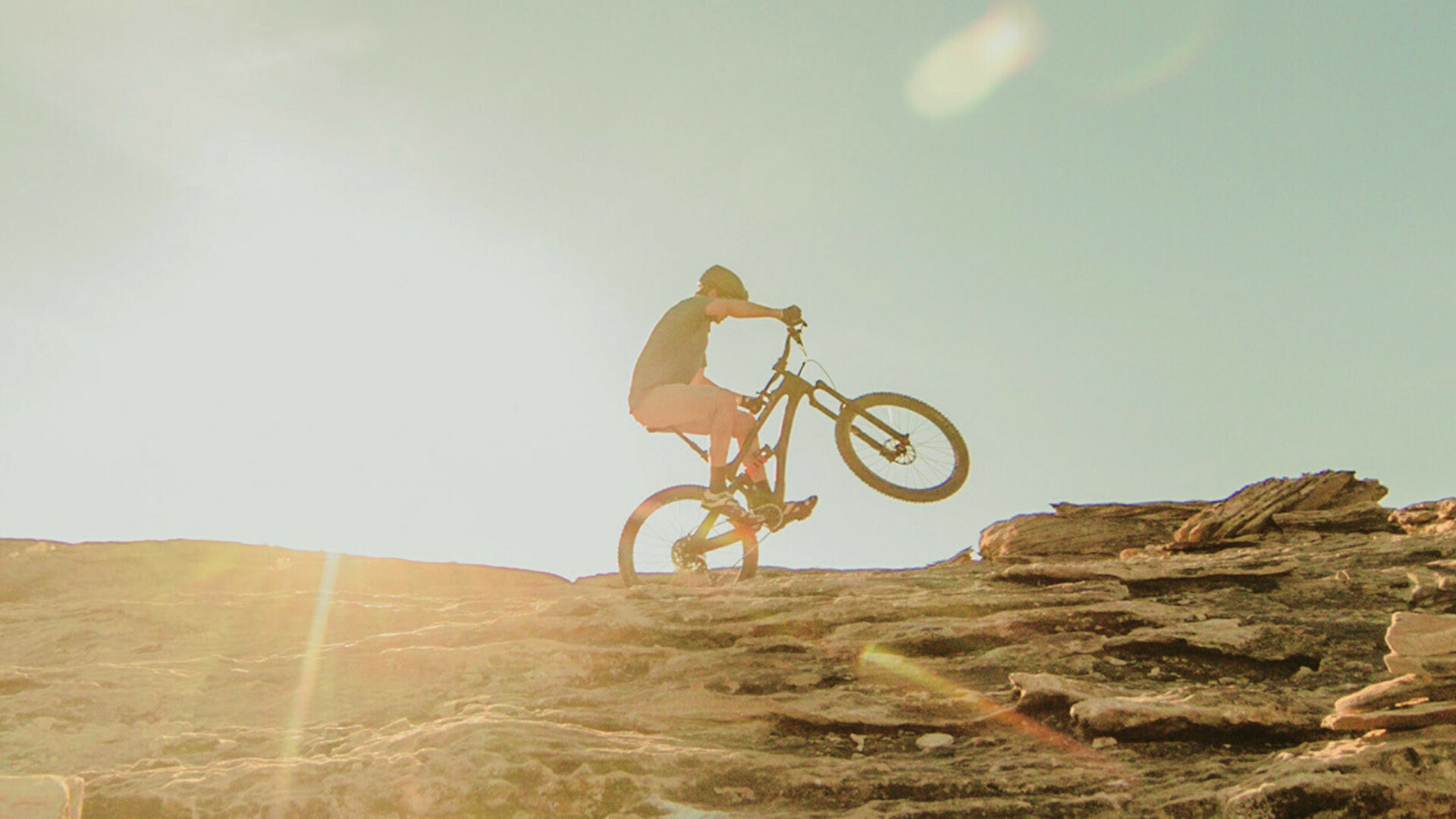 Mountain biker riding uphill on a rocky terrain during sunset, with sun rays visible in the background.