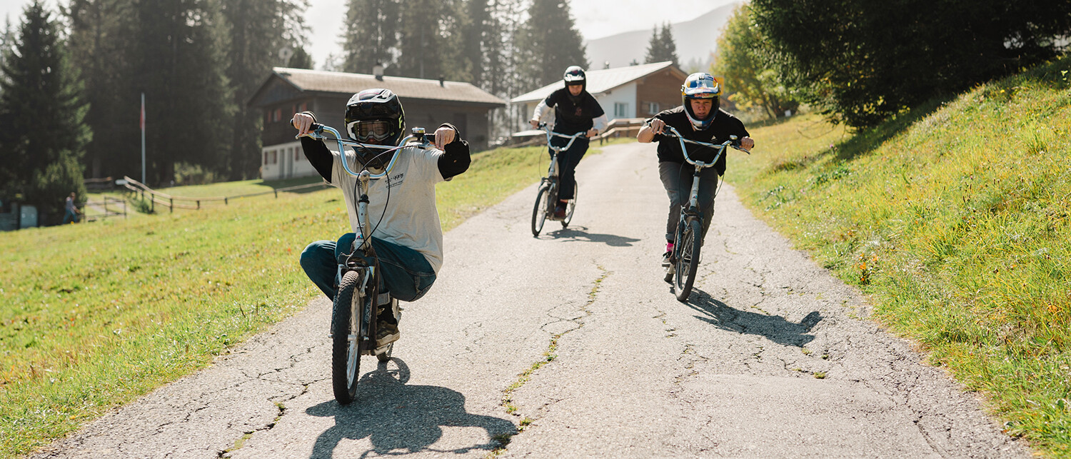 Trois cyclistes portant des casques descendent un chemin rural pavé, entourés d'herbe et d'arbres, avec des maisons en arrière-plan.