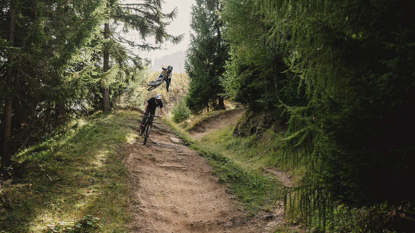 Un cycliste de montagne effectue une figure aérienne tandis qu'un autre cycliste navigue sur un sentier forestier. La scène est entourée de hauts arbres et d'une végétation luxuriante.