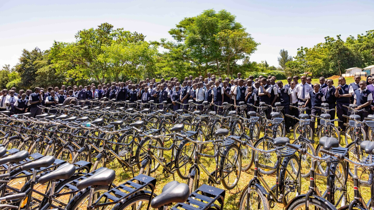 Un grand groupe d'élèves en uniforme se tient derrière une rangée de vélos empilés en plein air. Des arbres verts et un ciel dégagé sont visibles en arrière-plan.