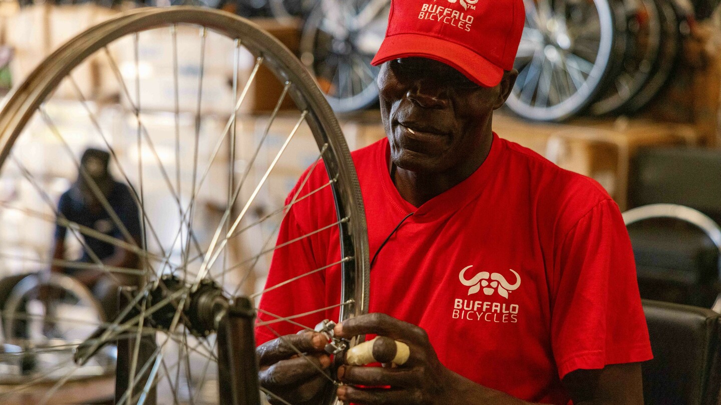 Un homme portant un t-shirt rouge et une casquette avec le logo Buffalo Bicycles travaille sur une roue de vélo dans un atelier. En arrière-plan, des vélos empilés et des cartons sont flous.