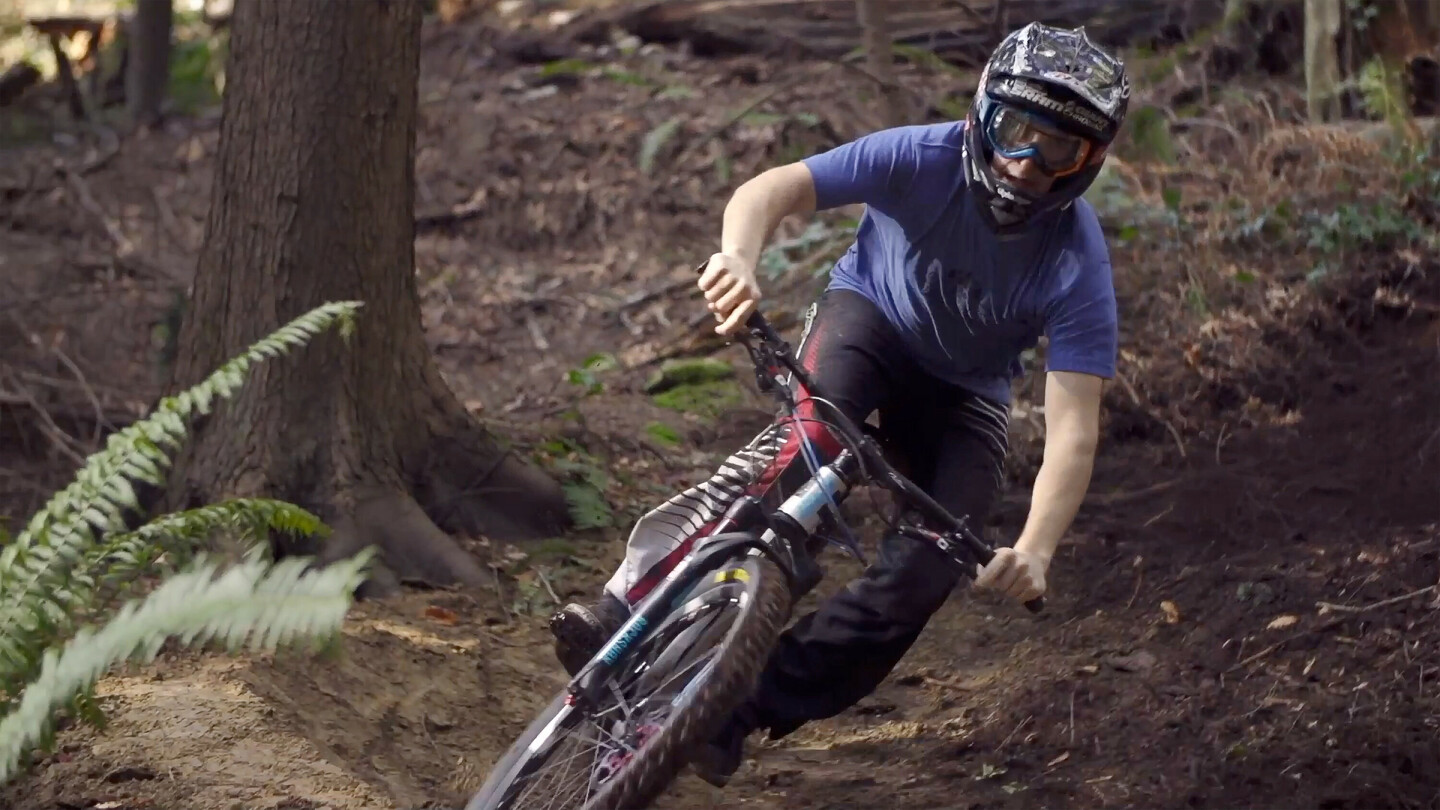 Un cycliste de montagne portant un casque et des lunettes traverse un virage dans une forêt. Le cycliste porte un t-shirt bleu et un pantalon noir, manœuvrant habilement le vélo sur un terrain inégal et terreux.