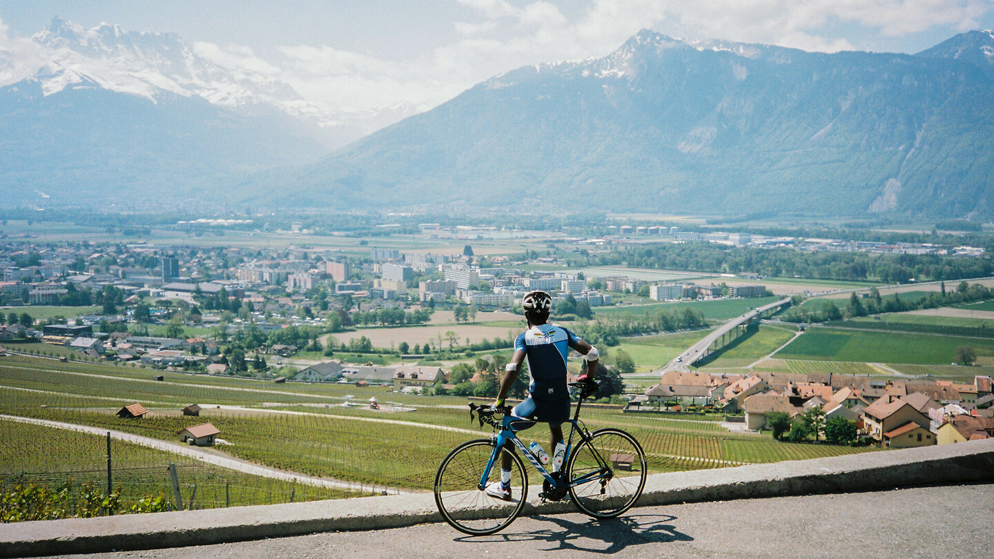 A cyclist standing next to their bike, overlooking a valley with a town, fields, and mountains in the background on a sunny day.