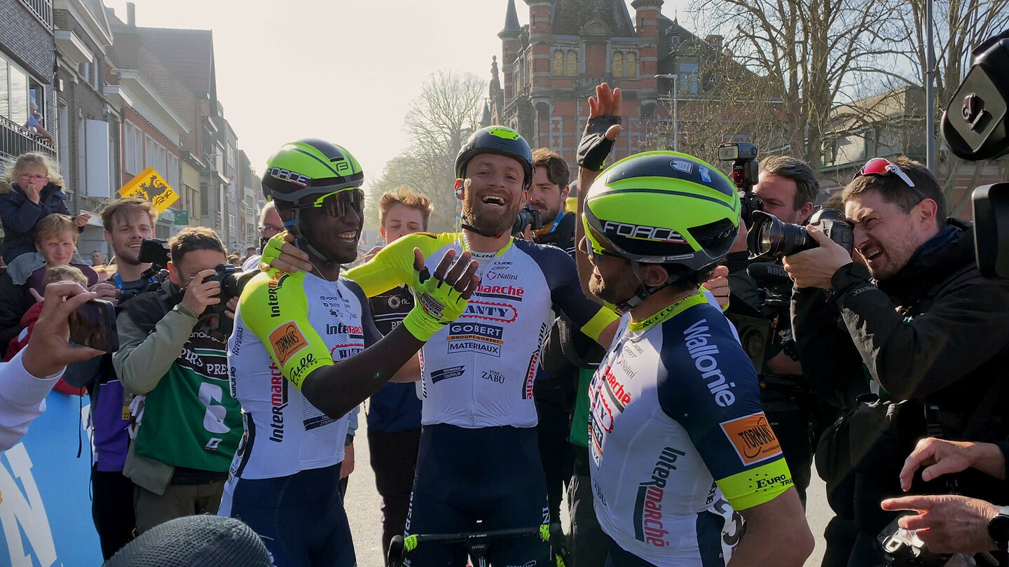 Road cyclists celebrating at the finish line surrounded by photographers and fans.