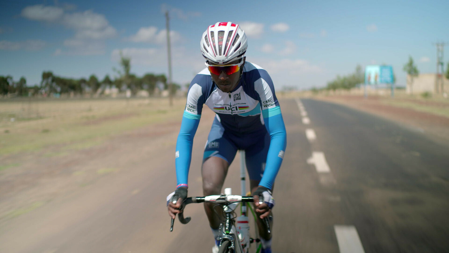 Biniam Girmay riding a road bike on an open road, wearing a blue and white UCI cycling kit and helmet.